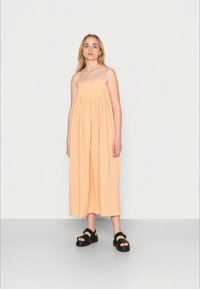 Jovem mulher em pé, vestindo um vestido pérola sem mangas e sandálias de plataforma pretas, contra um fundo claro e liso.