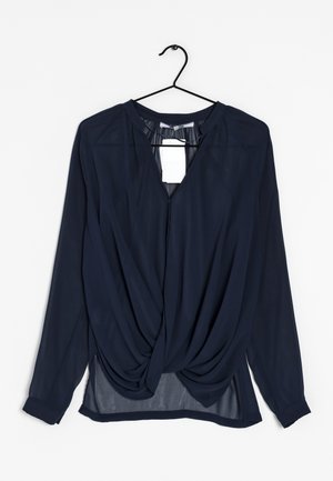 Blouse bleu marine à manches longues avec un design torsadé à l'avant suspendue sur un cintre noir sur fond blanc.