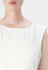 Hvit kjole med bred halslinning og plisserte detaljer, korte ermer og en jevn tekstur. Enkel og minimalistisk design.