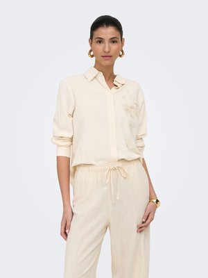 Femme portant une chemise crème à manches longues avec broderie florale et pantalon assorti à cordon, accessoirisée avec des boucles d'oreilles créoles en or et un bracelet.