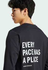 T-shirt noir à manches longues avec le texte blanc "CHAQUE RYTHME A UN ENDROIT" et "ADIDAS RUNNERS" dans le dos ; texture douce, design à col rond.