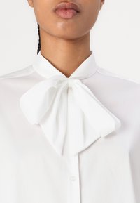 Blusa bianca con fiocco al collo, caratterizzata da una consistenza liscia e leggera e una chiusura con bottoni classica.