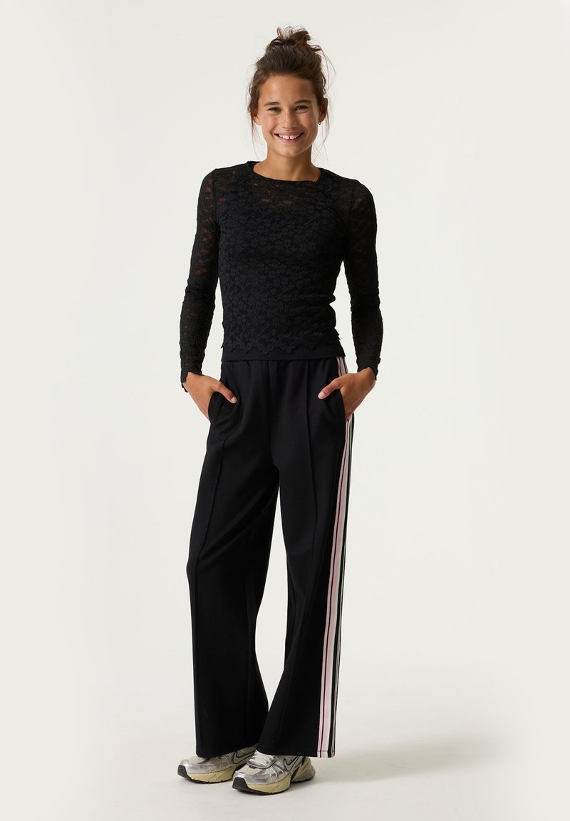 Schwarzes Spitzenoberteil mit langen Ärmeln und floralem Muster, kombiniert mit lockeren schwarzen Hosen mit pinkfarbener Seitenstreifen und Taschen, getragen mit silbernen Sneakers.