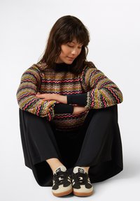 Mehrfarbiger Strickpulli mit wellenförmigen Mustern, schwarzen Bündchen, kombiniert mit schwarzen Palazzo-Hosen und grau-schwarzen Sneakers mit bernsteinfarbenen Akzenten.