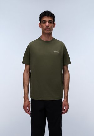 Bărbat stând purtând un tricou verde măsliniu cu mânecă scurtă și pantaloni negri, pe un fundal simplu gri deschis.
