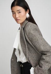 Καφέ και γκρι καμπαρντίνα με σχέδιο καρό, φορεμένη πάνω από ένα λευκό μπλουζάκι με μεγάλη φιόγκο, συμπληρωμένη με ασημένια κρεμάστα σκουλαρίκια.