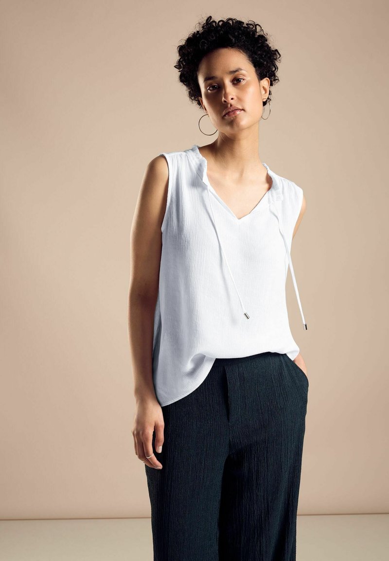 Blusa bianca senza maniche con scollatura a V e coulisse, abbinata a pantaloni scuri con texture. Tessuto morbido con vestibilità rilassata. Sfondo chiaro.