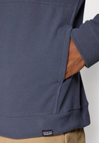 Fleece pullover i mørk marine med en blød, struktureret overflade. Har sidelommer og et synligt brandlabel nær kanten.