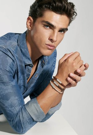 Blaue Jeanshemd mit hochgekrempelten Ärmeln, bronzefarbenen Knöpfen und drei Armbändern in Braun, Beige und Grau mit Textdetails. Glatte Haut, neutraler Hintergrund.