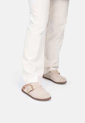 Zoccoli in suede beige con una ampia cinturino e dettaglio della fibbia dorata, poggiati su una suola marrone liscia, indossati con pantaloni bianchi.
