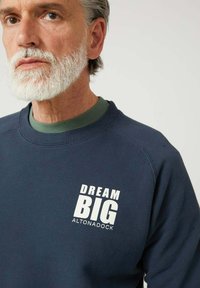 Hombre mayor con cabello y barba canosos, vistiendo un sudadera azul marino con el texto "DREAM BIG ALTONADOCK" en el pecho, fondo neutro.