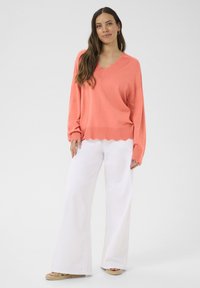 Koralen pulover z valovitim izrezom, prevelikim krojem in balonastimi rokavi. V kombinaciji z belimi širokimi hlačami in bež espadrilami.