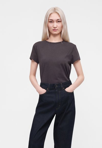 Femme aux longs cheveux blonds, portant un t-shirt sombre uni et un jean foncé taille haute, les mains dans les poches, debout devant un fond uni.
