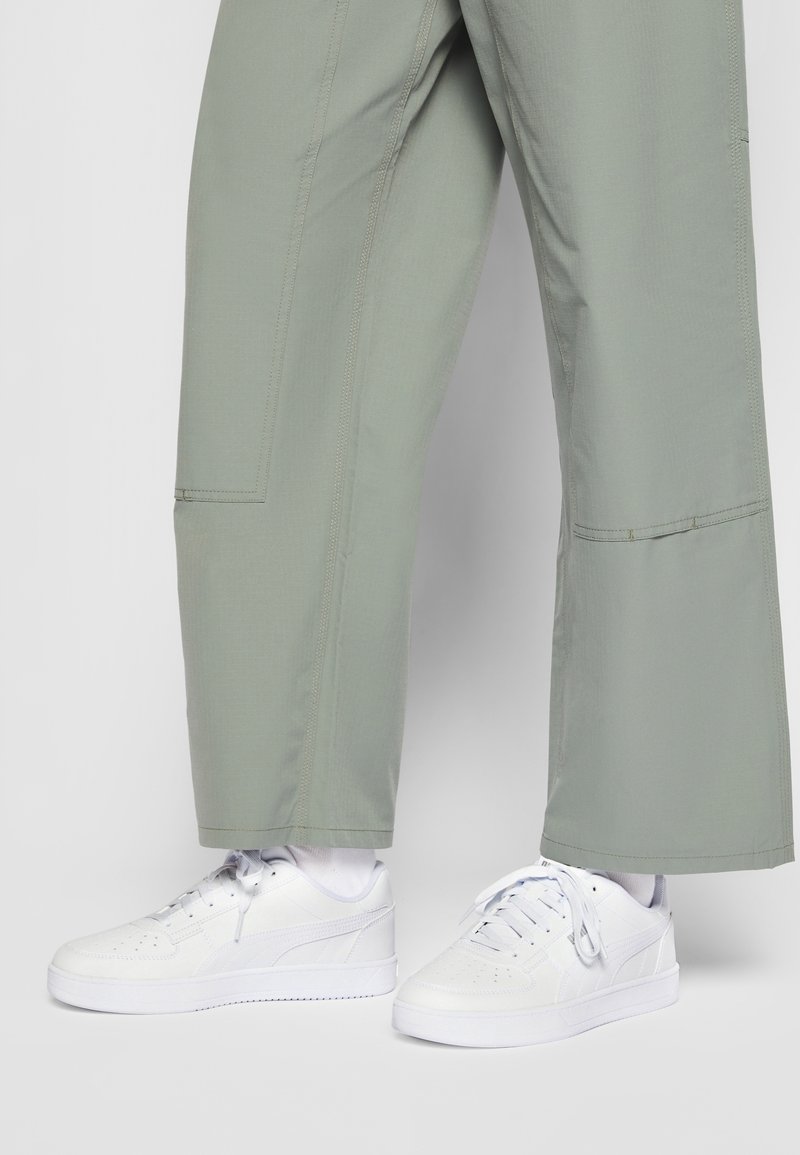 Jambes portant un pantalon large vert clair et des baskets blanches à lacets avec des chaussettes blanches sur un fond clair uni.