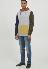 Kapuzensweatshirt mit grauem Körper, marineblauen und olivgrünen Ärmeln, gelber Fronttasche. Kombiniert mit blauen Jeans und schwarzen Sneakers.