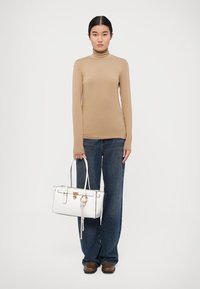 Bež turtleneck, tume sinised laiad teksad, pruunid kingad. Valge käekott kuldse riistvara ja dekoratiivse ahelaga. Üksne taust.