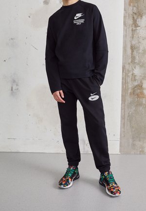 Person trägt schwarzen Nike-Sweatshirt mit dem Text "AUTHORIZED PERSONNEL ONLY", passende schwarze Nike-Sweatpants und bunte Turnschuhe.
