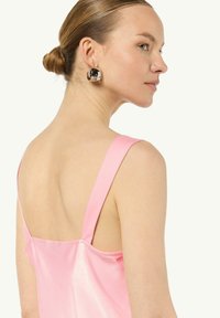 Różowa satynowa sukienka z szerokimi ramiączkami i niskim dekoltem z tyłu. Modelka w czarnych i srebrnych kolczykach-kołach, prezentująca gładką skórę i związane włosy.