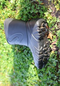 Zwarte rubberen laars met een gestructureerd oppervlak en diepe profielzool, gedeeltelijk ondergedompeld in groen gras en klaver.