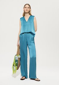 Türkises satiniertes ärmelloses Oberteil und weit geschnittene Hose mit lockerer Passform, kombiniert mit einer geflochtenen Tragetasche und einem floral gemusterten Schal.