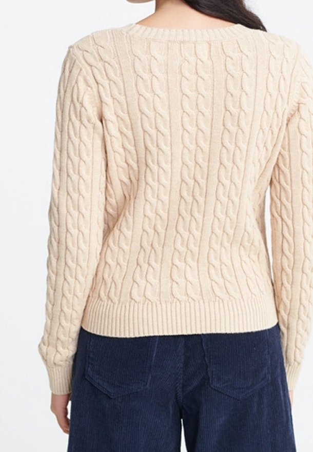 Pull beige en maille torsadée à manches longues porté par une personne debout, associé à un pantalon en velours côtelé bleu foncé.