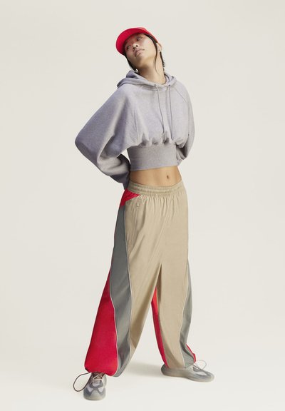 Persona que lleva una sudadera corta gris, pantalones anchos beige y rojos, zapatillas grises y una gorra roja se encuentra con las manos detrás de la espalda mirando hacia arriba.