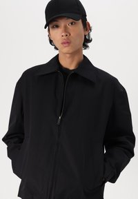 Črna zip-up jakna s koničastim ovratnikom, dvema sprednjima žepoma in gladko teksturo. Nosijo jo s črno kapo, ki se ujema.