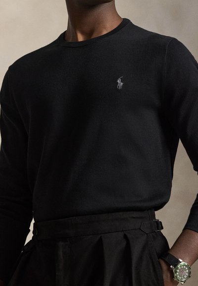 Person trägt ein schwarzes Langarmshirt mit kleinem besticktem Logo, schwarze Hose und eine Armbanduhr mit grünem Zifferblatt.