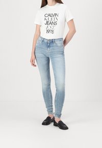 Mujer vistiendo una camiseta blanca de Calvin Klein Jeans y unos jeans ajustados azul claro, con mocasines negros, de pie contra un fondo blanco liso.