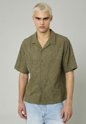 Chemise à manches courtes olive verte avec des motifs de feuilles en relief, col et boutons sur le devant. Coupe décontractée avec un tissu texturé.
