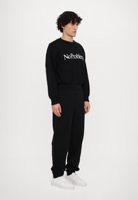 Schwarzer Sweatshirt mit "No Problem" in Weiß bedruckt, kombiniert mit schwarzen Jogginghosen. Das Modell trägt weiße Sneakers. Sanfter Stoff, lockere Passform.