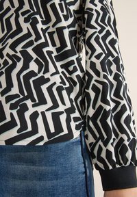Schwarz-weiß gemusterte Bluse mit abstrakten Formen, weicher Stoff, lange Ärmel mit elastischen Bündchen, kombiniert mit blauen Jeans.