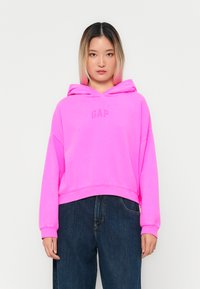 Rosa hettegenser med "GAP"-logo i en matchende nyanse, med cropped design og avslappet passform. Kombinert med mørk denimbukser.