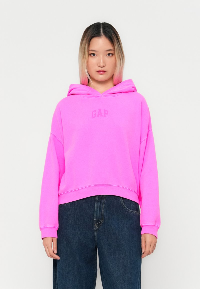 Rosa hettegenser med "GAP"-logo i en matchende nyanse, med cropped design og avslappet passform. Kombinert med mørk denimbukser.
