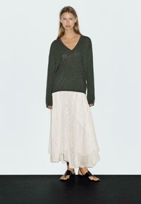 Dunkelgrüner Strickpullover mit V-Ausschnitt, kombiniert mit einem weißen, fließenden Langrock bis zum Knöchel mit einem Rautenmuster. Schwarze flache Sandalen.