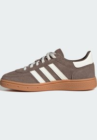 Brauner Wildleder-Sneaker in niedriger Ausführung mit weißen Streifen, weißem Fersenriemen, weißen Schnürsenkeln und strukturierter Sohle aus Kaugummirubber, seitlich dargestellt.