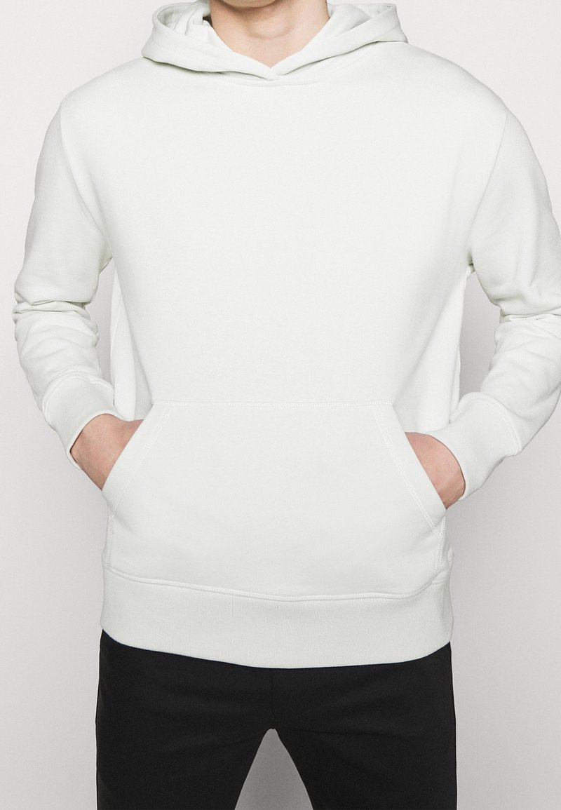 Sweat à capuche en coton blanc avec une poche kangourou avant, des poignets côtelés et une capuche réglable avec cordon de serrage. Texture lisse, design décontracté.