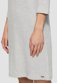 Graues, strukturiertes Kleid mit einem Wabenmuster und Dreiviertelärmeln, mit einem kleinen Markenakzent nahe dem Saum.
