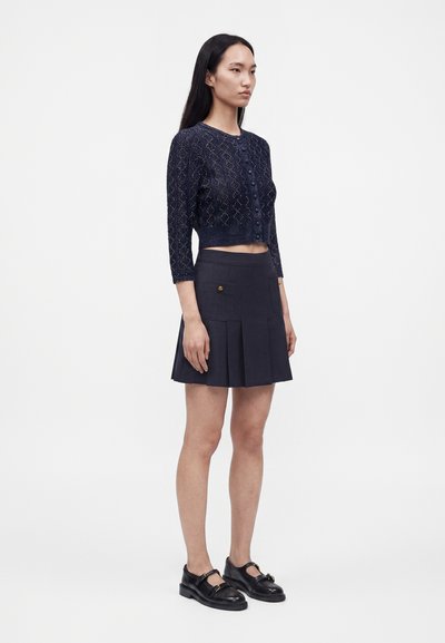 Femme debout portant un cardigan court en maille bleu marine, une jupe plissée bleu marine avec poches et des chaussures Mary Jane noires sur un fond uni.