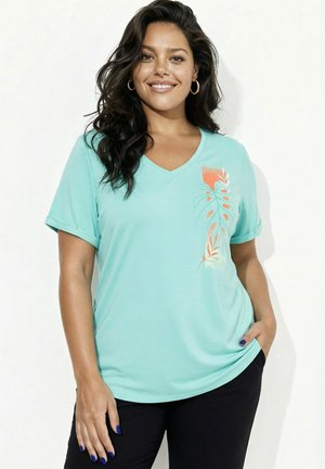 Uśmiechnięta kobieta z długimi ciemnymi włosami nosi jasnoniebieską koszulkę z dekoltem w serek z pomarańczowo-białym wzorem liści, czarne spodnie i złote kolczyki koła.