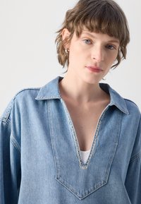 Camicetta in denim blu chiaro con scollo a V, maniche lunghe e colletto testurizzato. Presenta cuciture a contrasto lungo ilcollo e la patta.