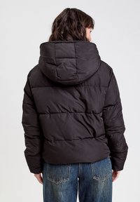 Doudoune noire avec capuche, présentant un design matelassé et une coupe décontractée, associée à un jean en denim bleu clair.