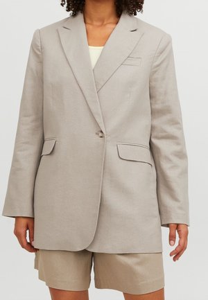 Femme portant un blazer beige à un seul bouton sur un top blanc et un short beige assorti, debout devant un fond uni.