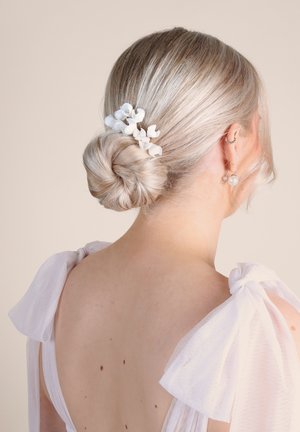 Haare zu einem niedrigen Dutt frisiert, geschmückt mit kleinen weißen Blumenclips. Das Haar ist hellblond mit glatter Textur, kombiniert mit einem transparenten weißen Trägerkleid.