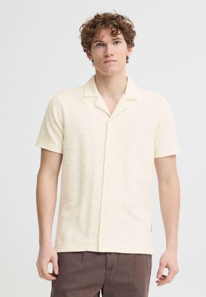 Ung mand med krøllet hår iført en cremefarvet kortærmet skjorte med knapper og brune bukser, stående foran en enkel hvid baggrund.