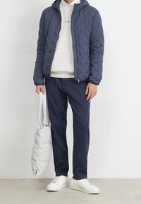 Man draagt een marineblauwe gewatteerde jas, crème sweatshirt, marineblauwe broek, witte sneakers, houdt een witte gewatteerde draagtas vast, staand op een beige vloer.