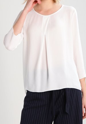 Frau trägt eine locker sitzende weiße Bluse mit 3/4-Ärmeln und marineblaue, weit geschnittene Hose mit Nadelstreifen, posiert mit einer Hand nahe dem Gesicht.