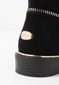 Sort ruskind støvle med en tyk sort sål, der har et guldaccentbånd, en guldlogo-plade og en dekorativ lynlåsdetalje.