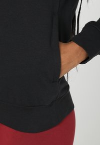 Felpa nera con una texture liscia, dotata di tasca laterale e maniche a costine, abbinata a pantaloni rossi aderenti.