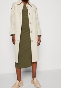 Persona que lleva un abrigo largo beige sobre un vestido midi verde oliva de manga larga y mocasines beige gruesos.